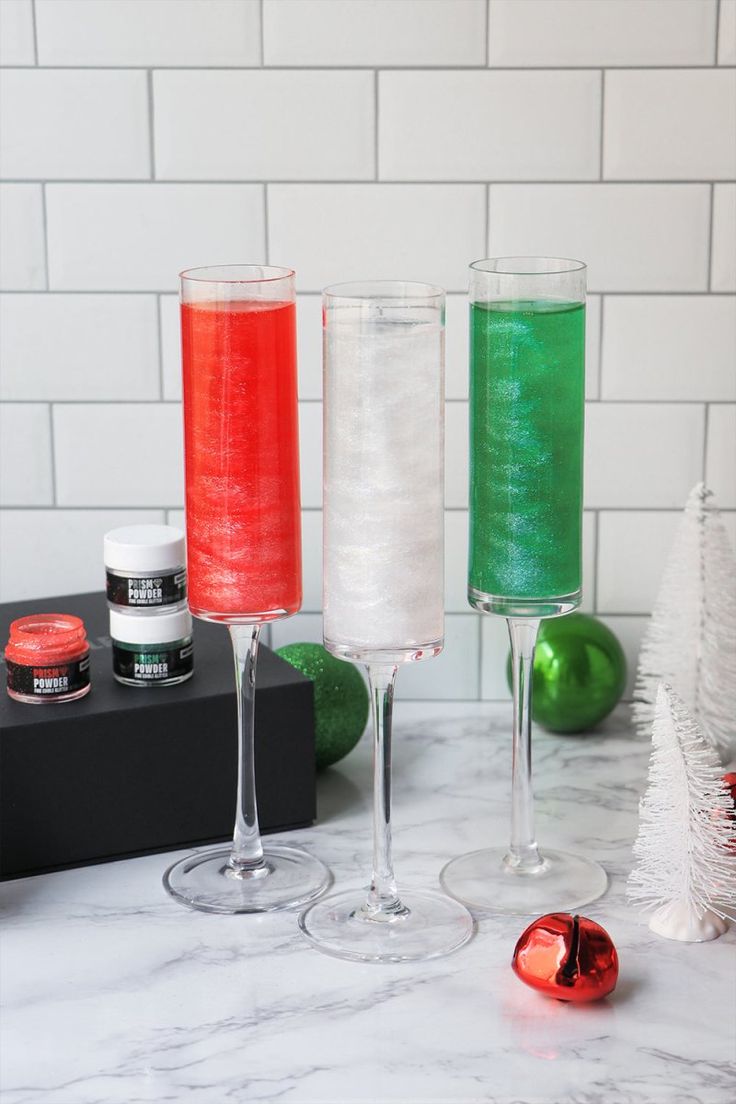 three wine glasses filled with colored liquid on top of a white marble counter next to christmas decorations