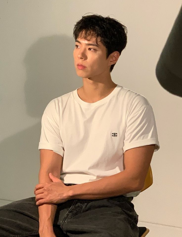 a young man sitting on top of a wooden chair next to a lamp and wearing a white t - shirt