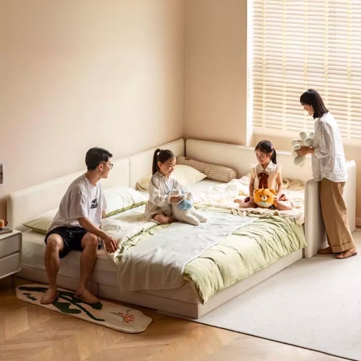 three people sitting on a bed in a room with white walls and wooden flooring