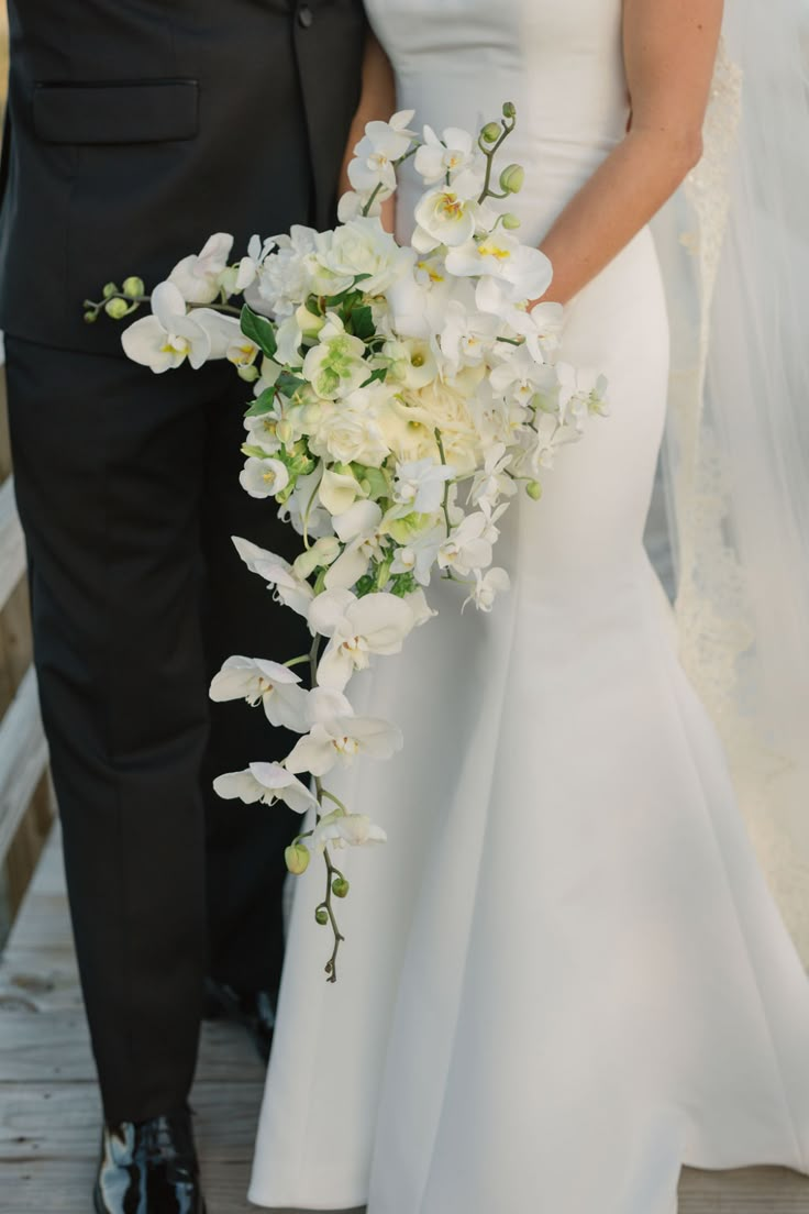 a bride and groom standing on a dock with flowers in their hands, dressed in black tuxedo