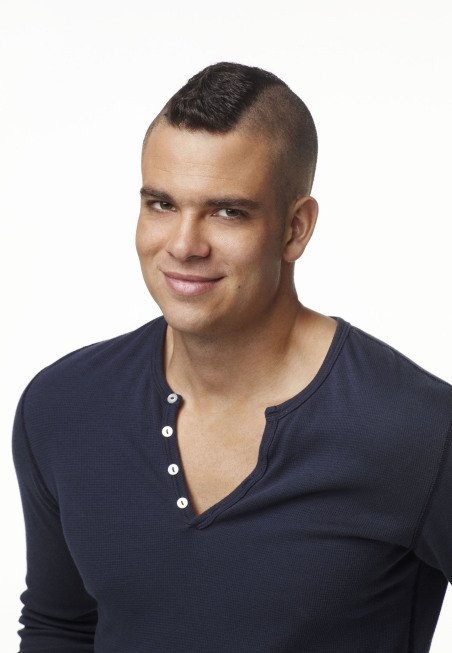 a man with a shaved head wearing a blue shirt and smiling at the camera while standing in front of a white background