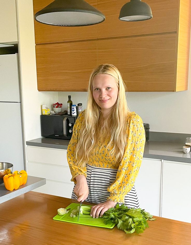 a woman standing in a kitchen cutting vegetables on a green cutting board and smiling at the camera