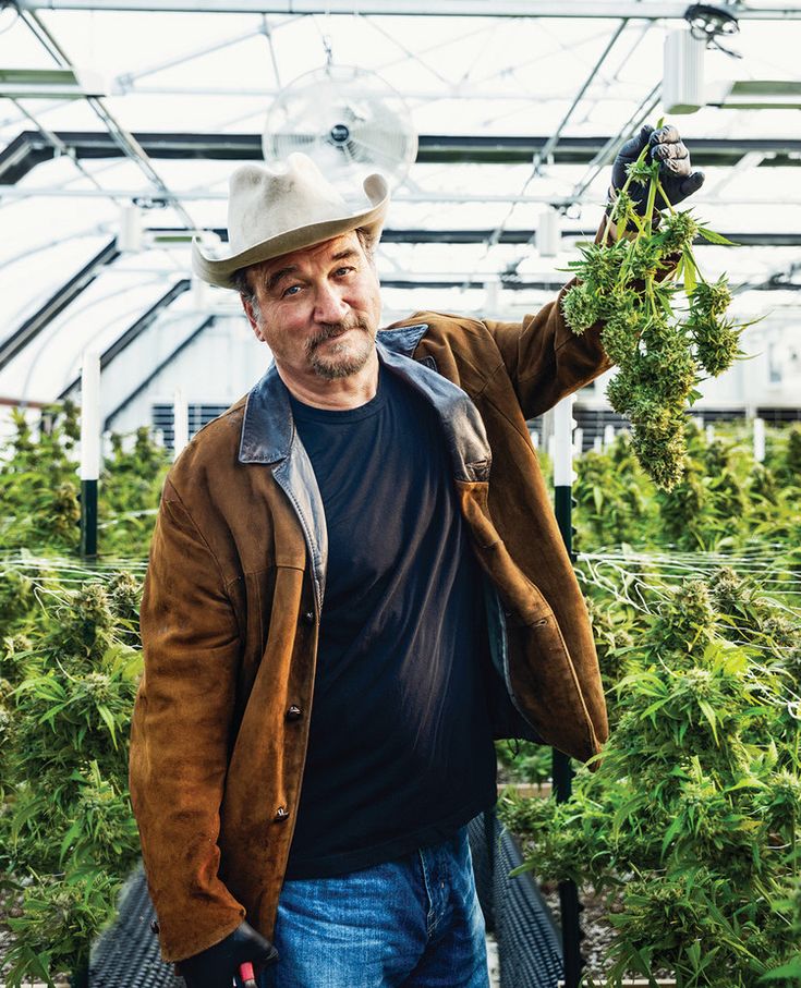 a man wearing a hat and holding up some plants in a greenhouse with other plants behind him