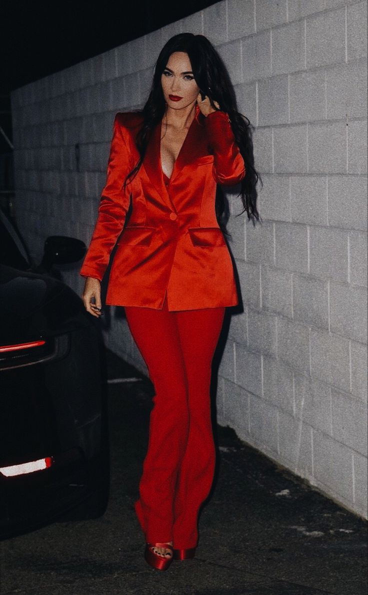 a woman in a red suit is standing next to a white wall and talking on her cell phone