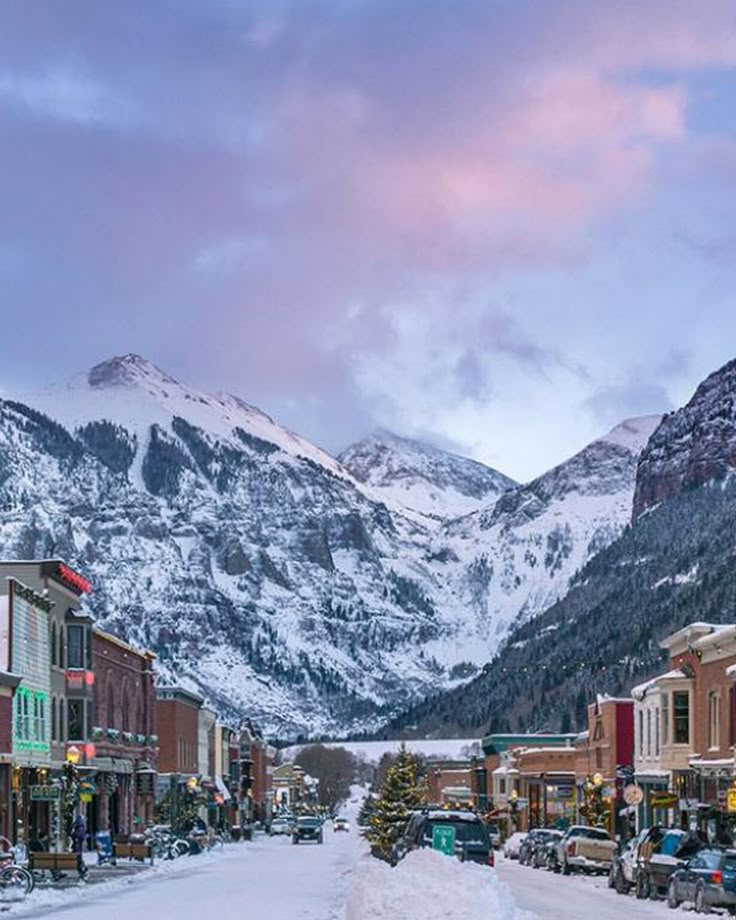 a snowy street lined with buildings and mountains in the background at dusk or dawn,