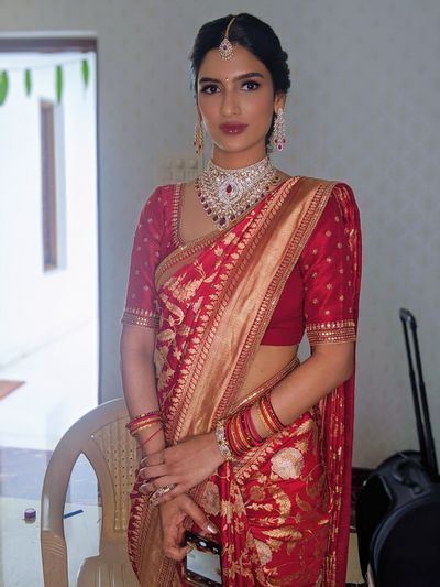 a woman in a red and gold sari with jewelry on her neck, standing next to a chair