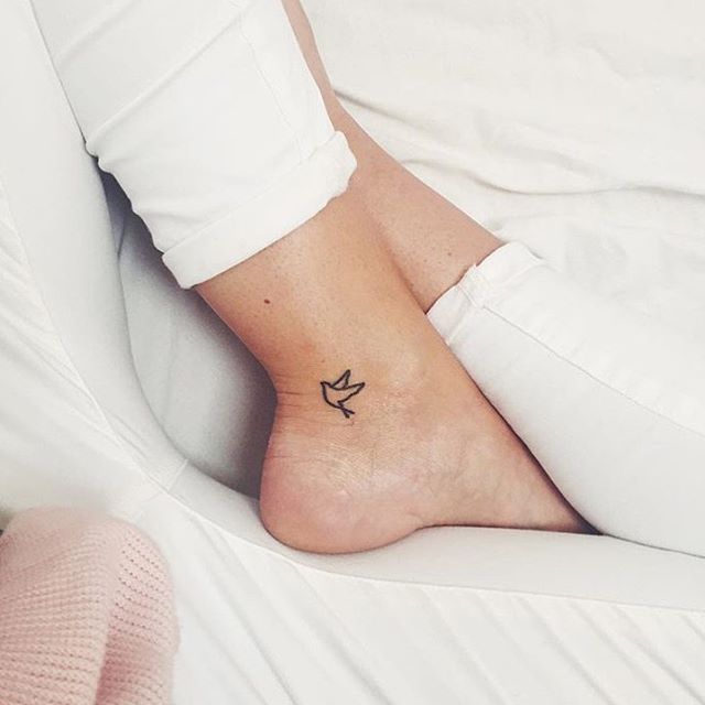 a person with a small bird tattoo on their foot sitting on a white bed next to a pink blanket