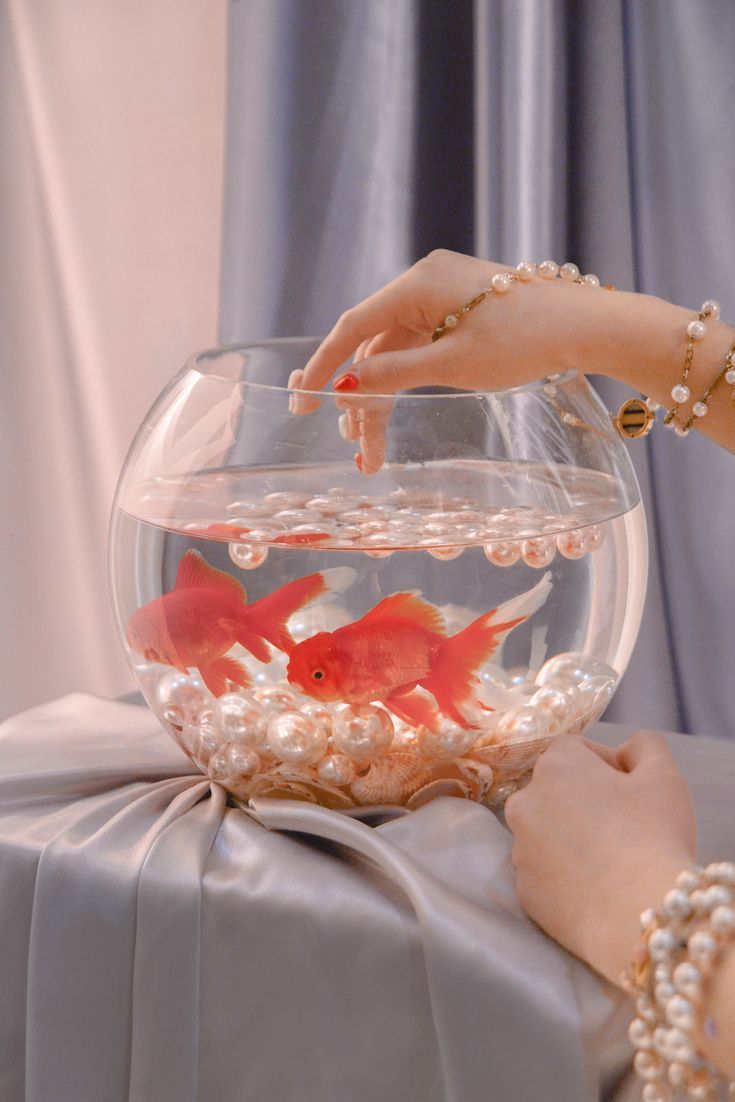 a woman's hand reaching for a goldfish in a bowl with pearls on it