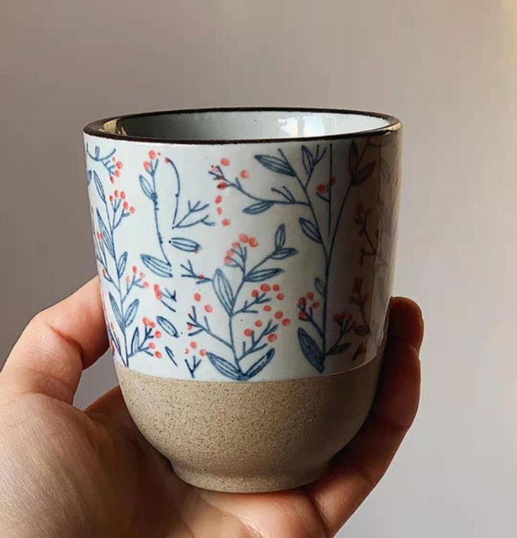 a hand holding a blue and white cup with red berries on the outside, in front of a gray background