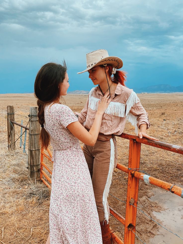 two women standing next to each other in front of a fence with an open field behind them