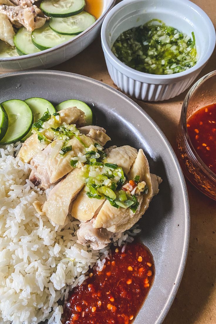 On a grey plate is a serving of rice topped with chicken, ginger scallion sauce, spicy sauce, and sliced cucumbers. Next to the plate is an extra serving of ginger scallion sauce in a ramekin and extra serving of spicy sauce in a clear bowl. Hainese Chicken Rice, Singapore Chicken Rice Recipe, Hainese Chicken, Chicken Rice Easy, Ginger Rice Recipe, Singaporean Rice, Singapore Chicken Rice, Hainan Chicken Rice, Easy Vietnamese Recipes