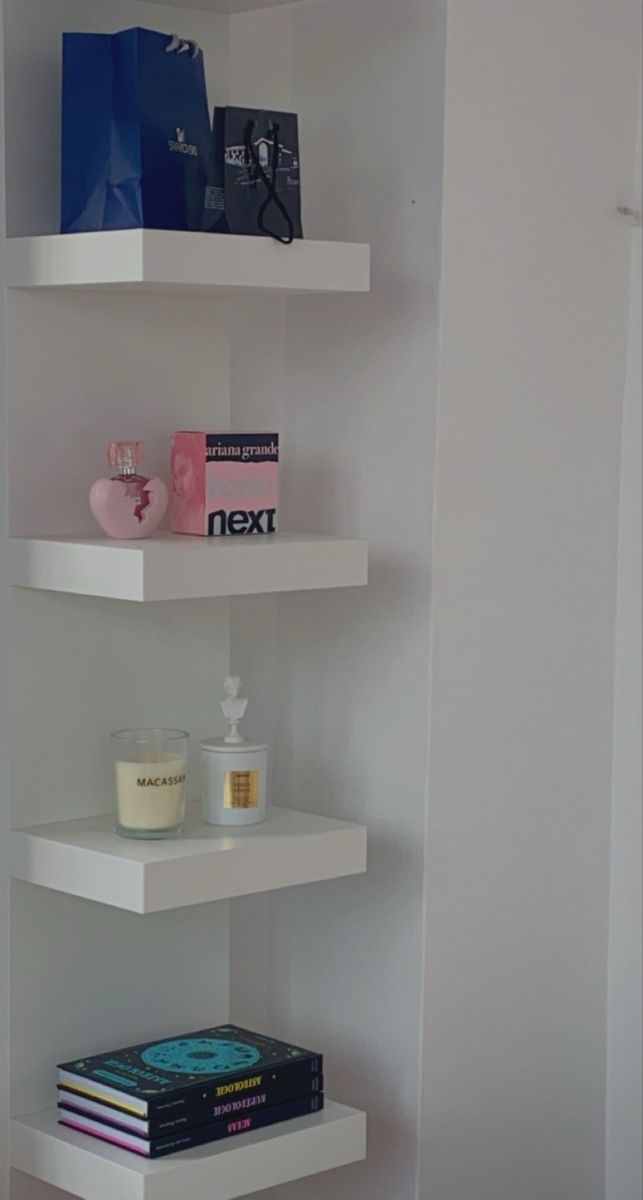 three white shelves with books, candles and other items on them in a corner room