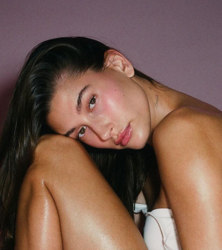 a beautiful young woman sitting on top of a bed next to a purple wall with her hands behind her head