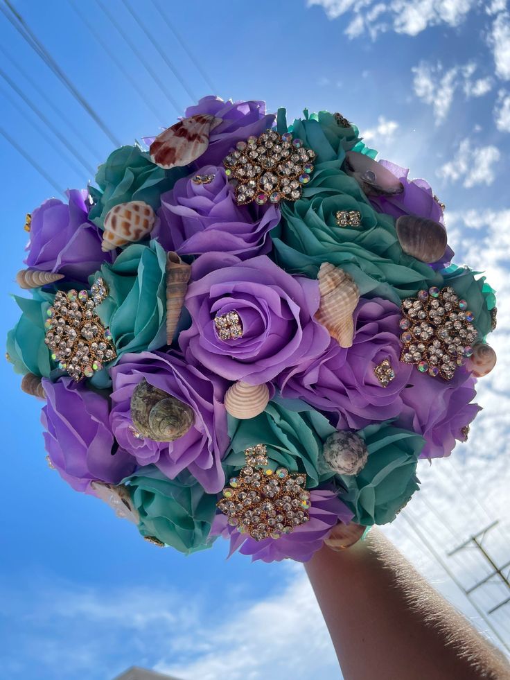 a bridal bouquet made with purple flowers and seashells is held up in the air