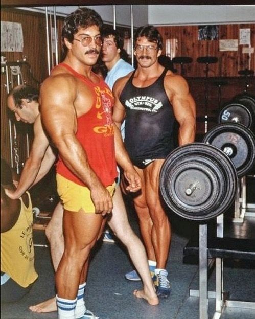 two men standing next to each other in front of a barbell rack with one man showing off his muscles