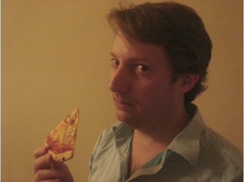 a man holding up a slice of pizza in his right hand and looking at the camera