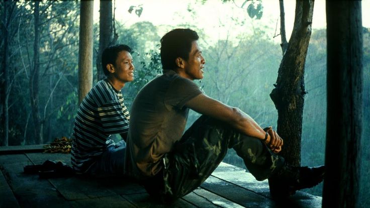 two men sitting on a wooden deck in the woods looking at each other and smiling