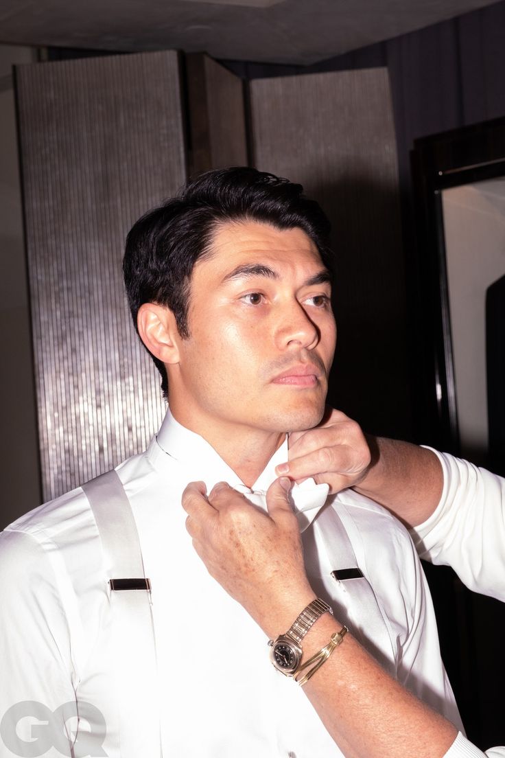 a man adjusting his tie while standing in front of a mirror wearing a white shirt and suspenders