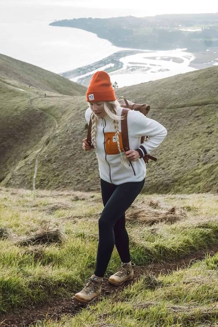 a woman wearing an orange hat and hiking gear on top of a grassy hill next to the ocean