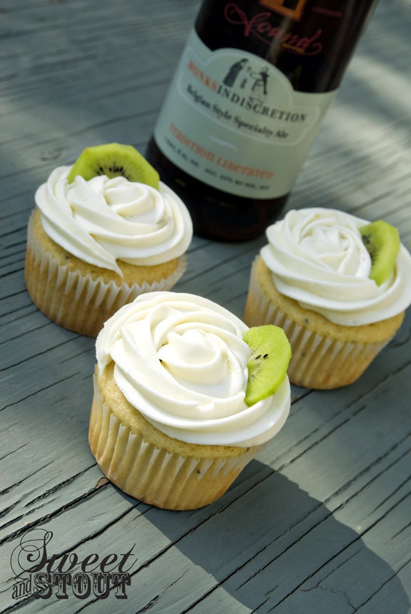 three cupcakes with white frosting and kiwi slices on them next to a bottle of wine