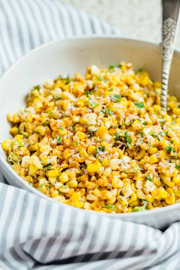 a white bowl filled with corn on top of a blue and white striped cloth next to a spoon