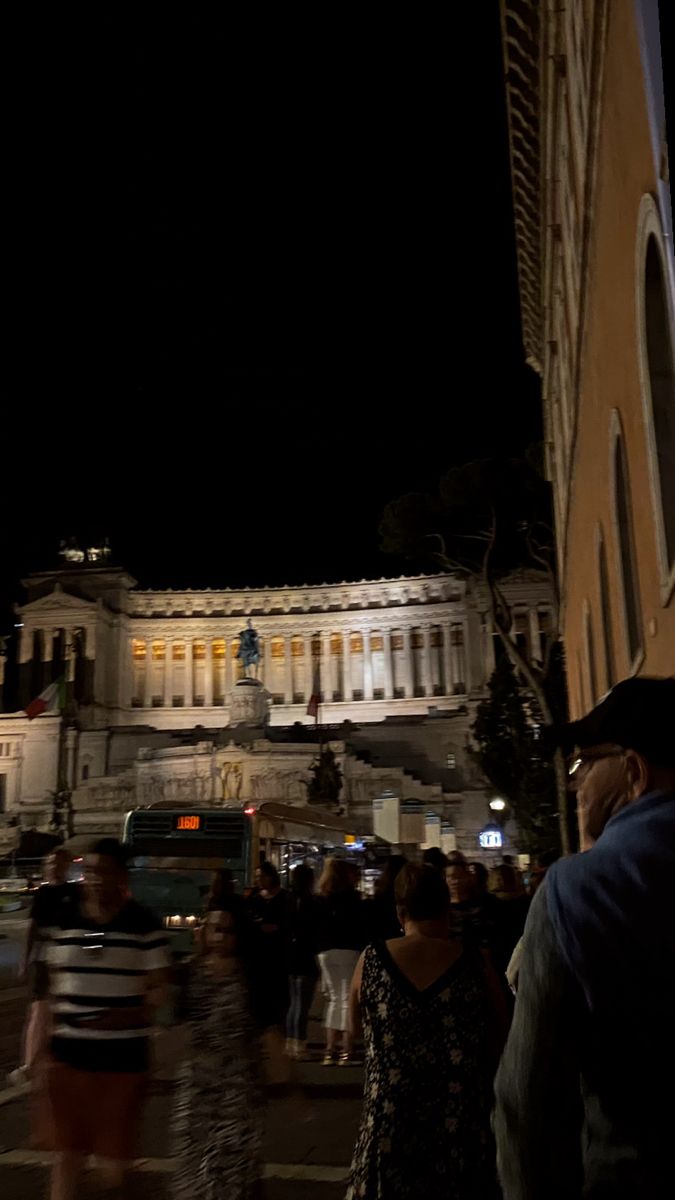 people are walking down the street in front of a large building at night with lights on