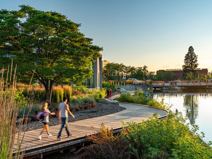 two people walking across a wooden bridge over a river next to tall grass and trees