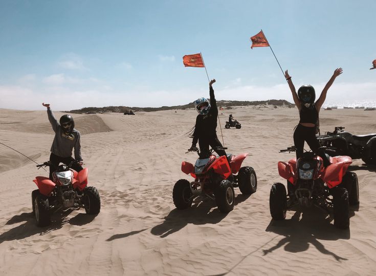 three people on four wheelers in the sand with flags and two atv's