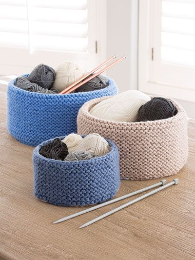 three knitted baskets with yarn and knitting needles on a table in front of windows