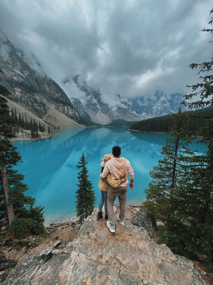 two people standing on the edge of a cliff looking at a blue lake with mountains in the background