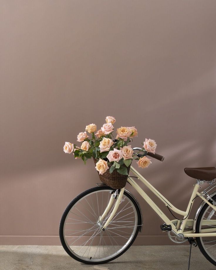 a bicycle with flowers in the basket parked next to a pink wall and wooden chair