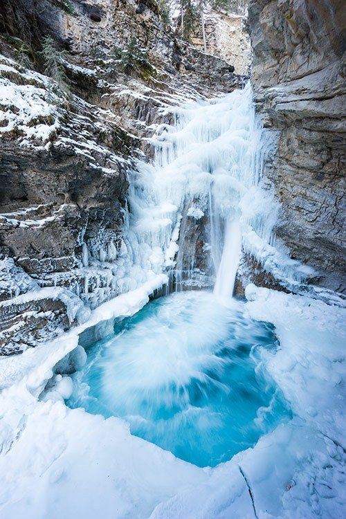 a frozen waterfall in the middle of a snowy mountain area with blue water running down it's sides