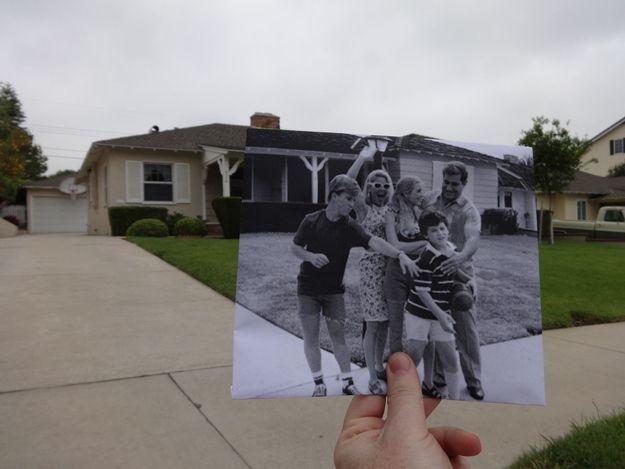 a person holding up an old photo in front of a house with the family on it