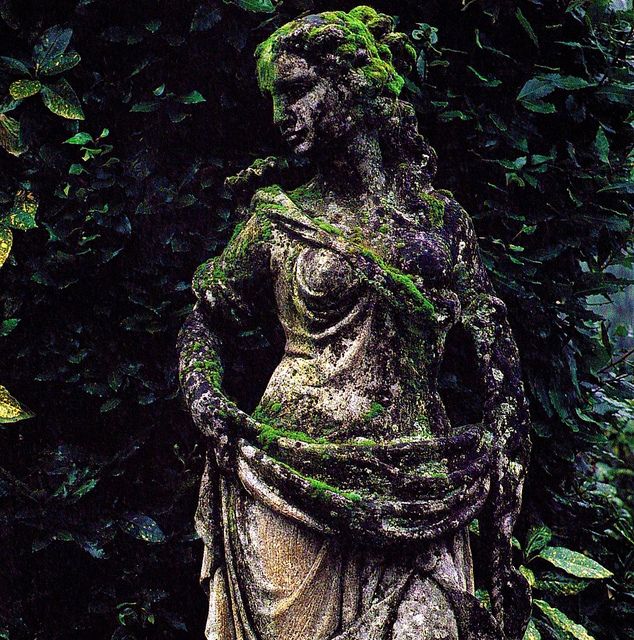 a statue with moss growing on it in front of some trees and bushes, surrounded by greenery