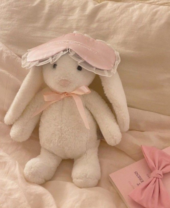 a white stuffed rabbit with a pink hat on it's head sitting next to a book