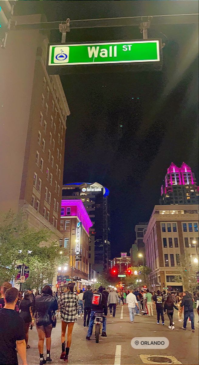 people are walking down the street in front of some tall buildings at night, and there is a green sign that says wall st