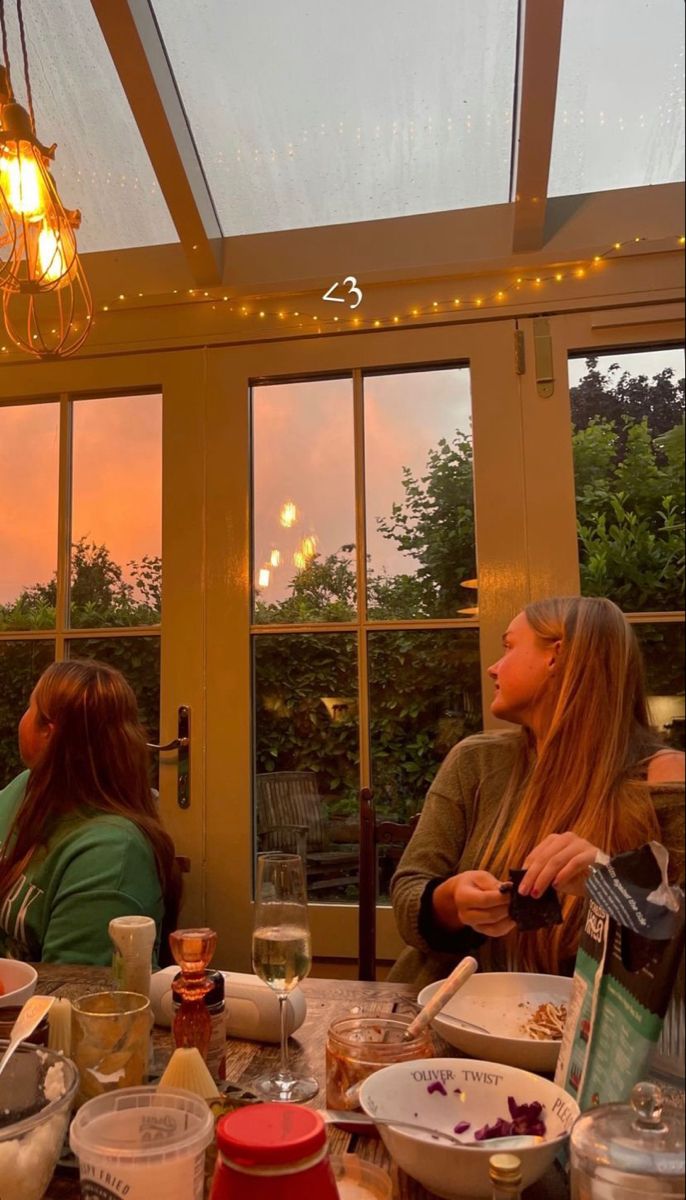 two women sitting at a table with food and drinks in front of them, looking out the window