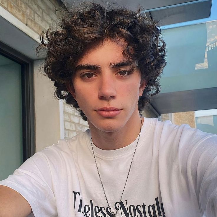 a young man with curly hair wearing a white t - shirt and looking at the camera