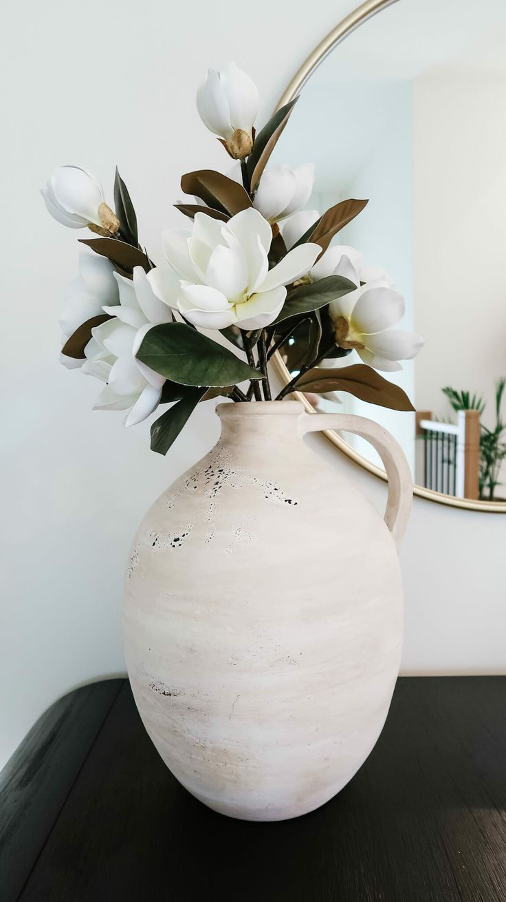 a white vase with flowers in it sitting on a wooden table next to a mirror