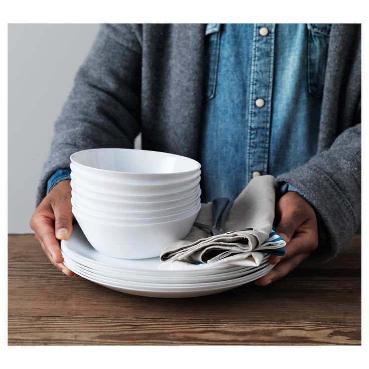 a person holding a stack of white plates on top of a wooden table with silverware in their hands