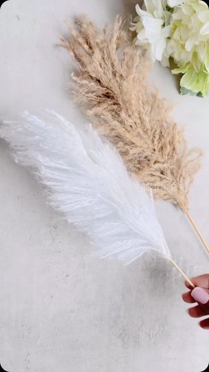 a hand holding a feather on top of a white table next to flowers and a vase