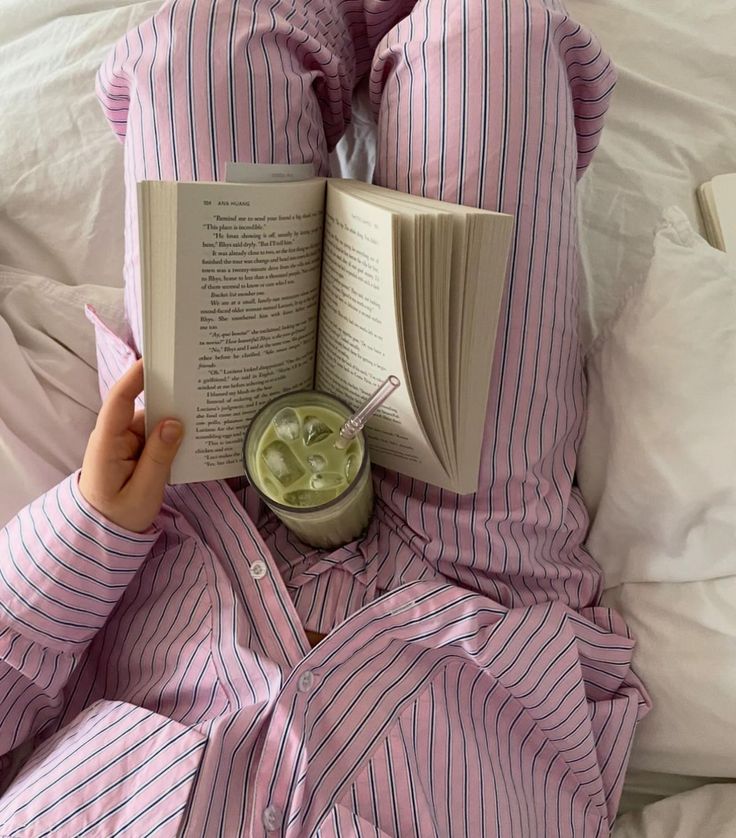 a person laying in bed reading a book and drinking green liquid from a cup on top of an open book