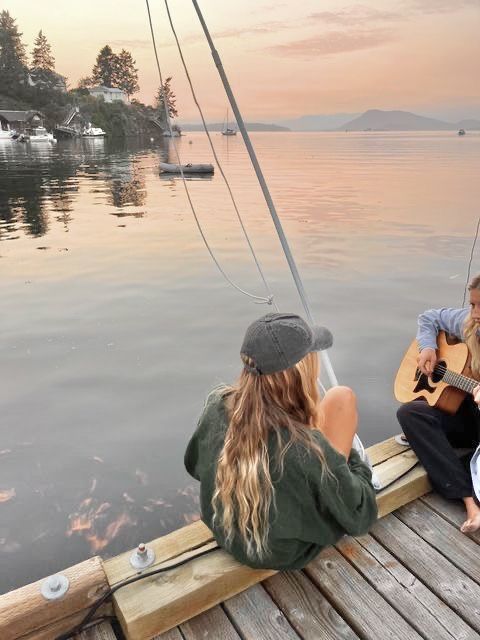 two women are sitting on a dock playing the guitar and another woman is looking at them