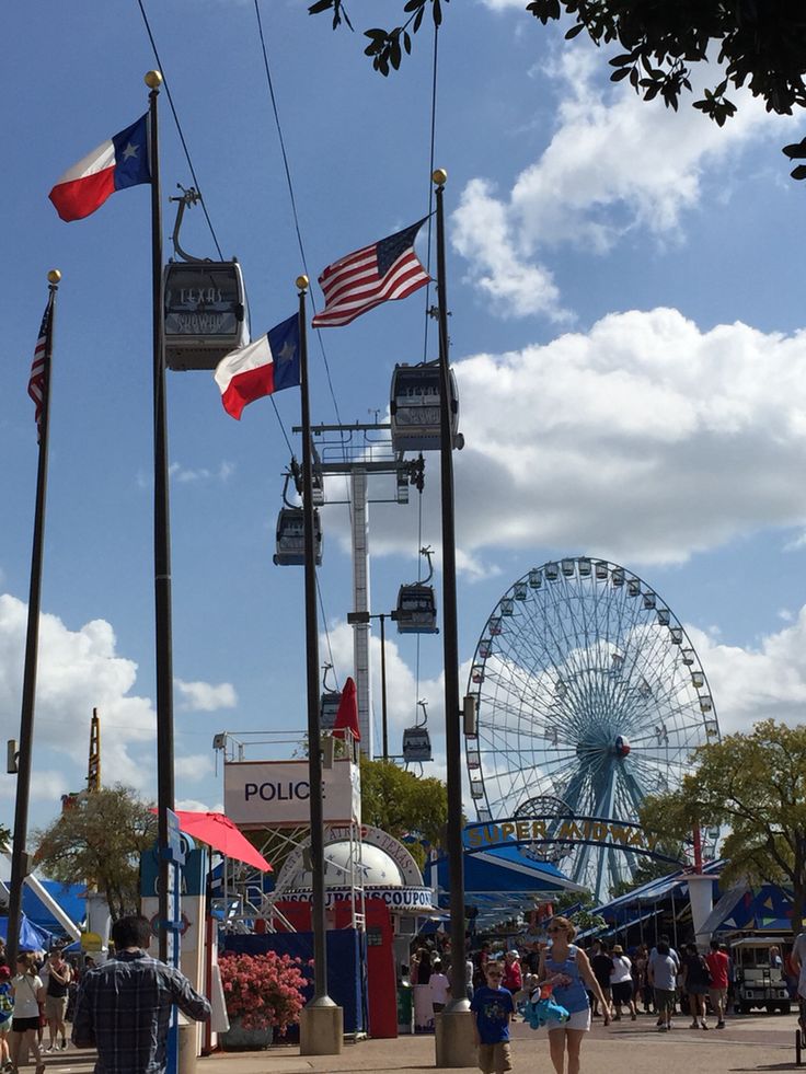 people walking around an amusement park with flags and ferris wheel in the background on a sunny day