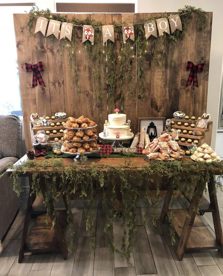 a table topped with lots of pastries next to a wooden wall covered in greenery