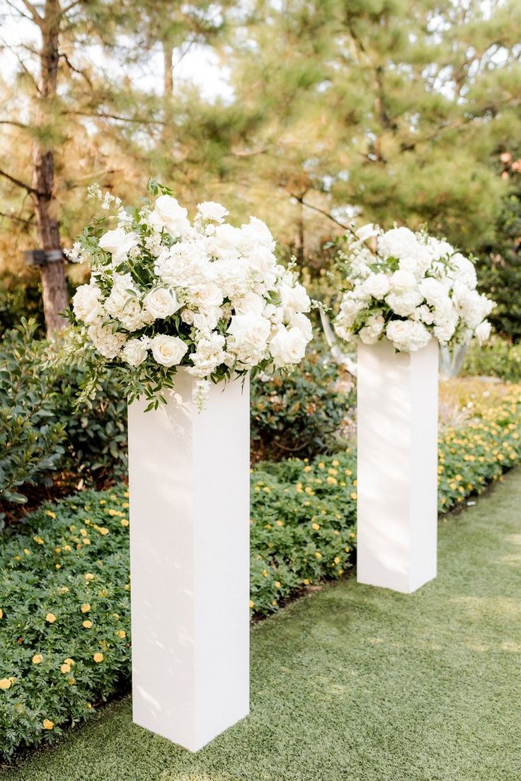 two tall vases with white flowers are on the grass near some bushes and trees