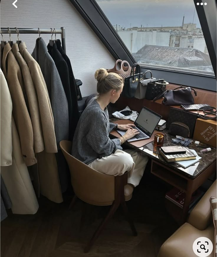 a woman sitting at a desk in front of a window working on a laptop computer