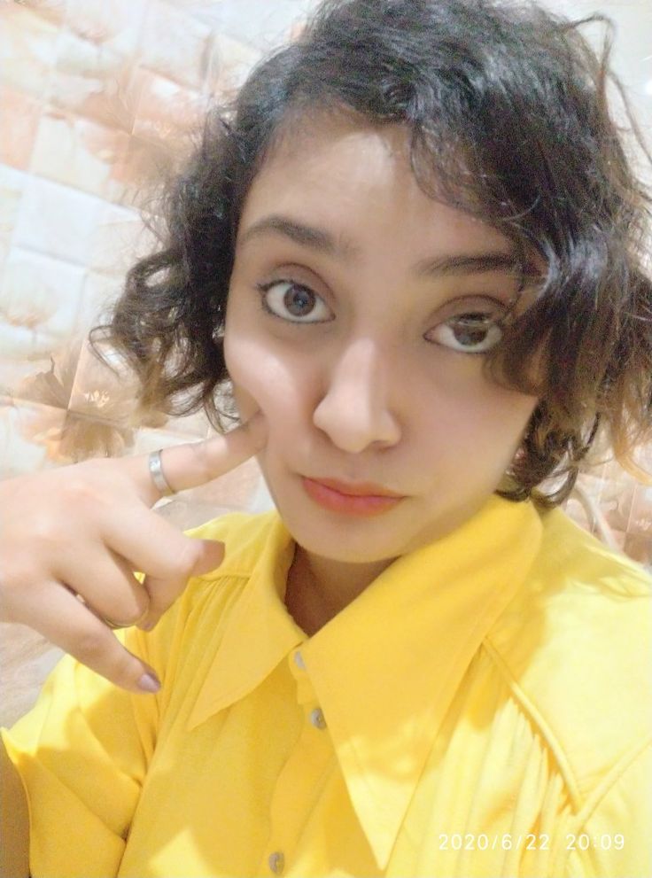 a woman with curly hair wearing a yellow shirt and pointing to her left side while looking at the camera