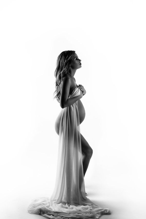 black and white photograph of pregnant woman in flowing dress, standing on one leg with her hands behind her back