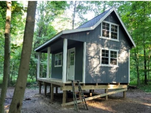 a small cabin in the woods with stairs leading up to it's door and windows